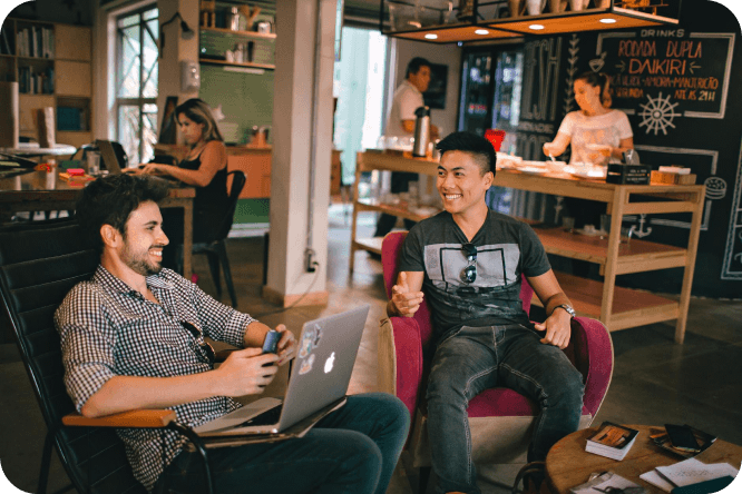 Two people sitting in a office, engaged in a friendly conversation with laptops and notebooks on the table.